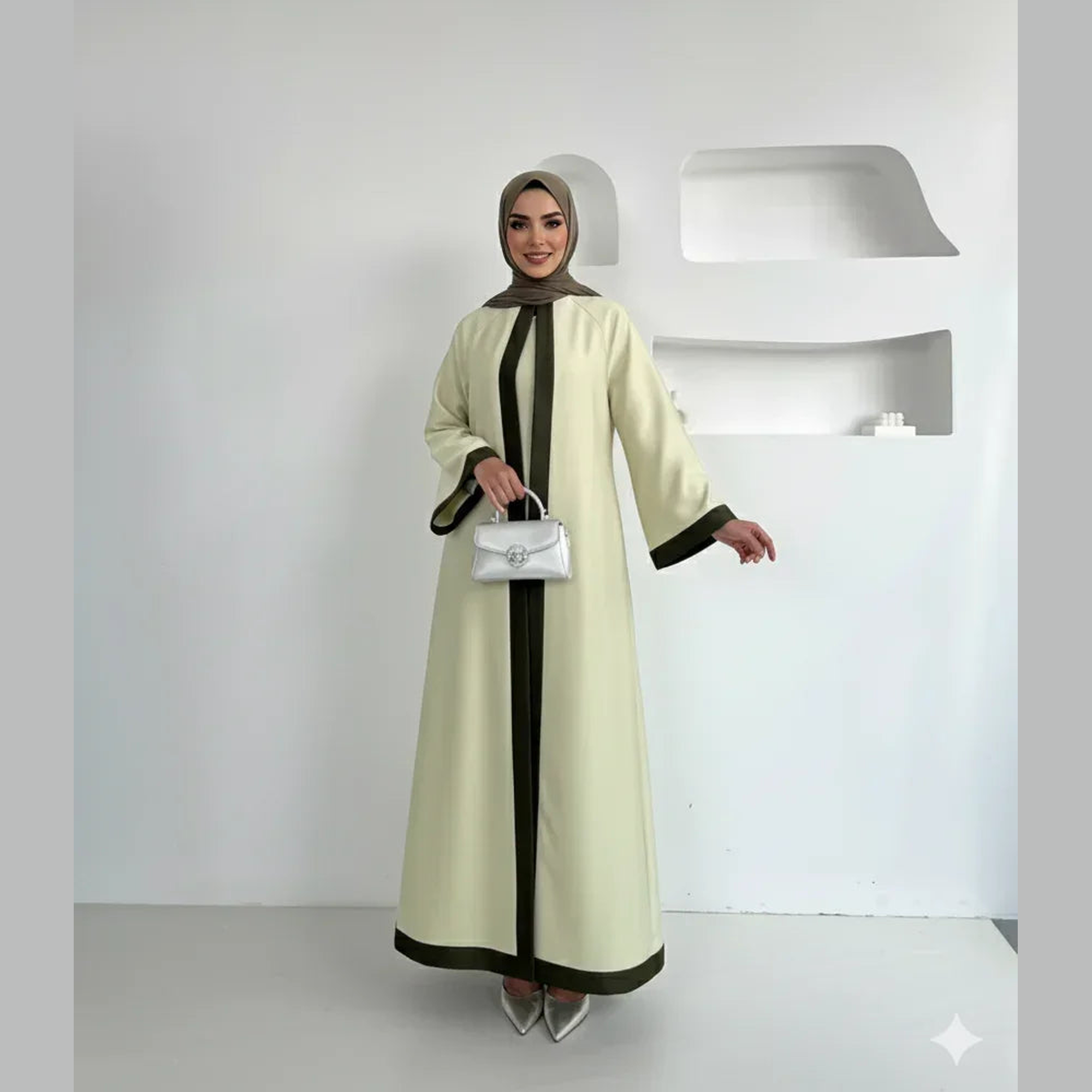 Woman in elegant beige abaya with black trim, taupe hijab, and silver purse, standing in modern minimalist room. Modest fashion from ZAHRA'S BEST.
