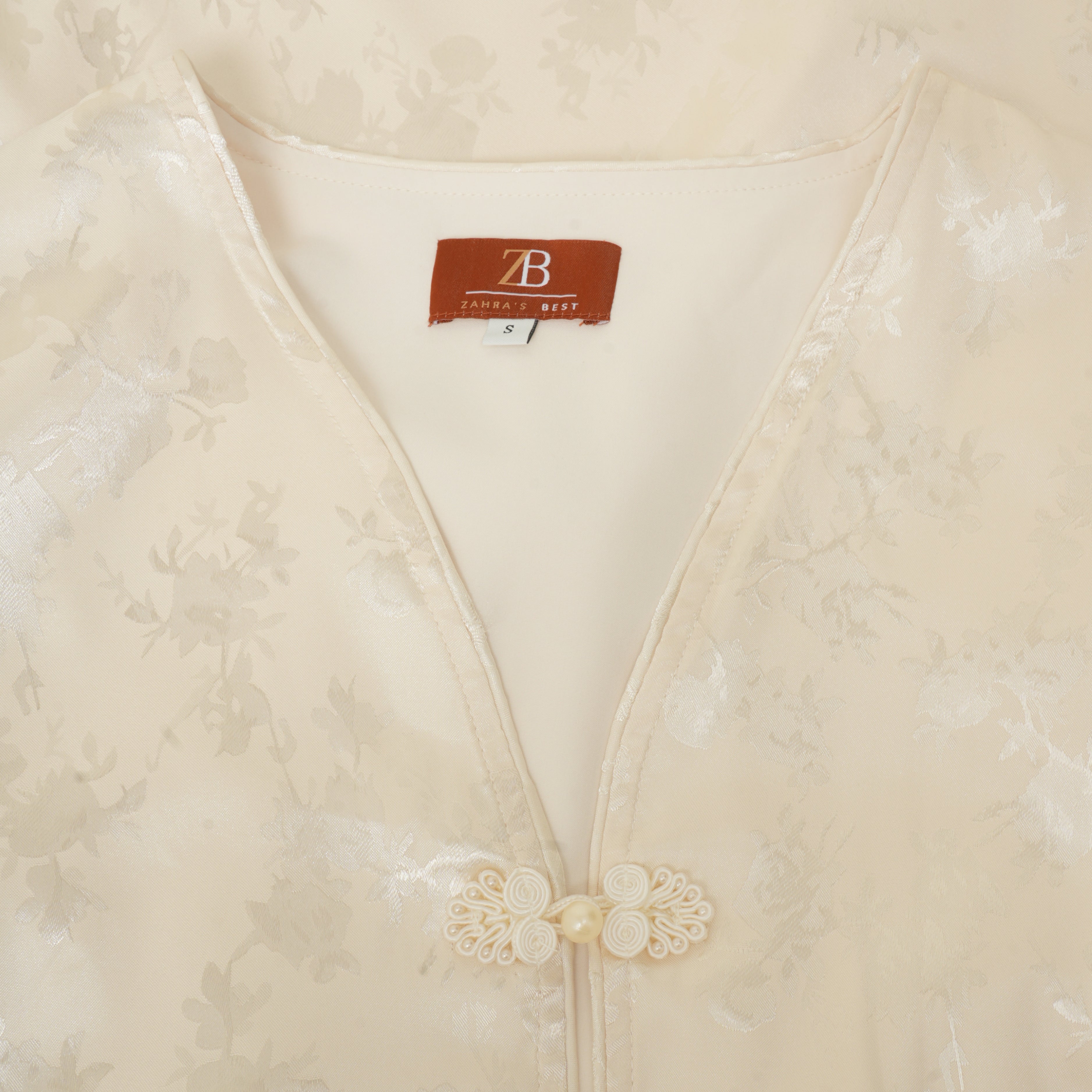 Close-up of a cream-colored Chinese-style blouse with a subtle floral jacquard pattern, featuring a V-neckline and decorative lace-and-pearl button closure. The inner neck label shows the brand 'B.