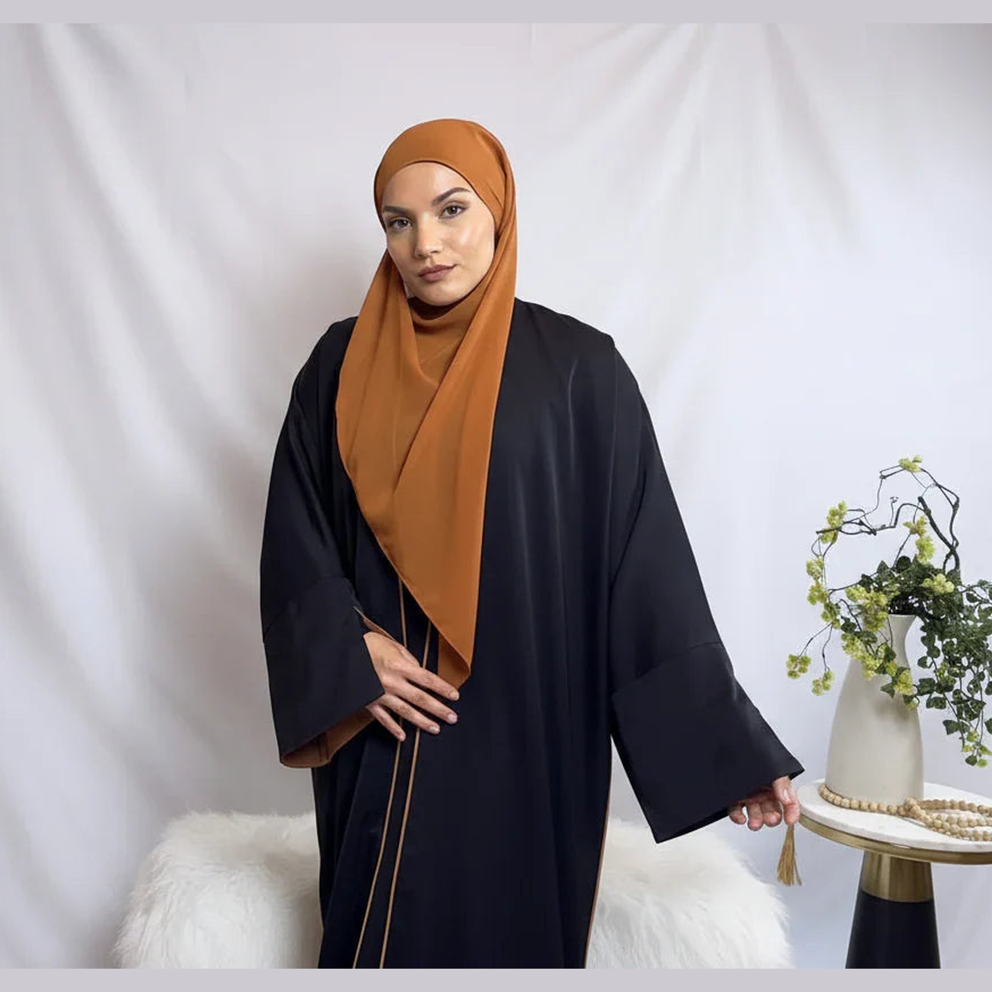 Woman in a brown hijab and black abaya stands by a table with a vase, showcasing modest fashion