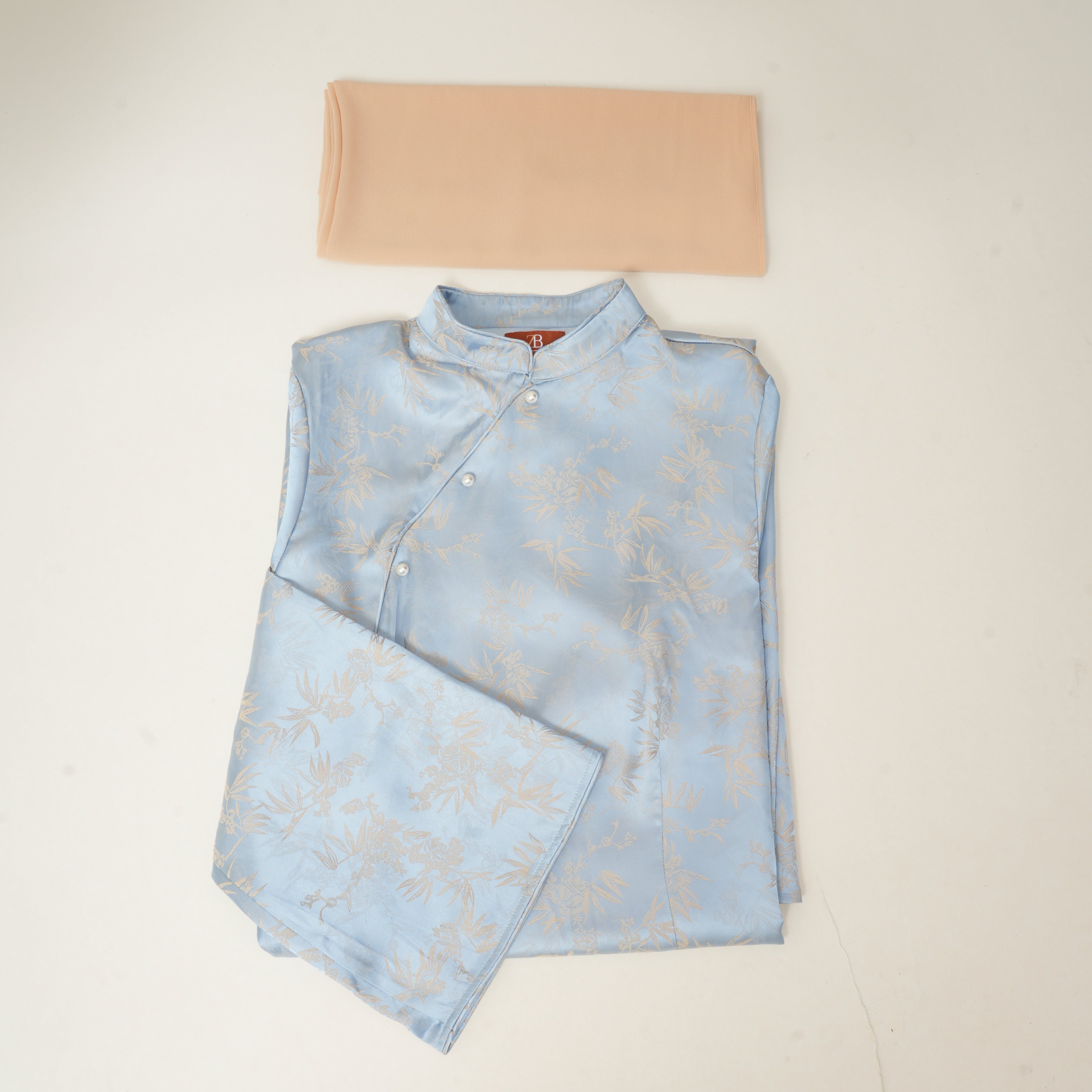 A folded light blue satin-like garment with a subtle beige floral pattern, shown with a matching beige neck wrap on a plain off-white background.