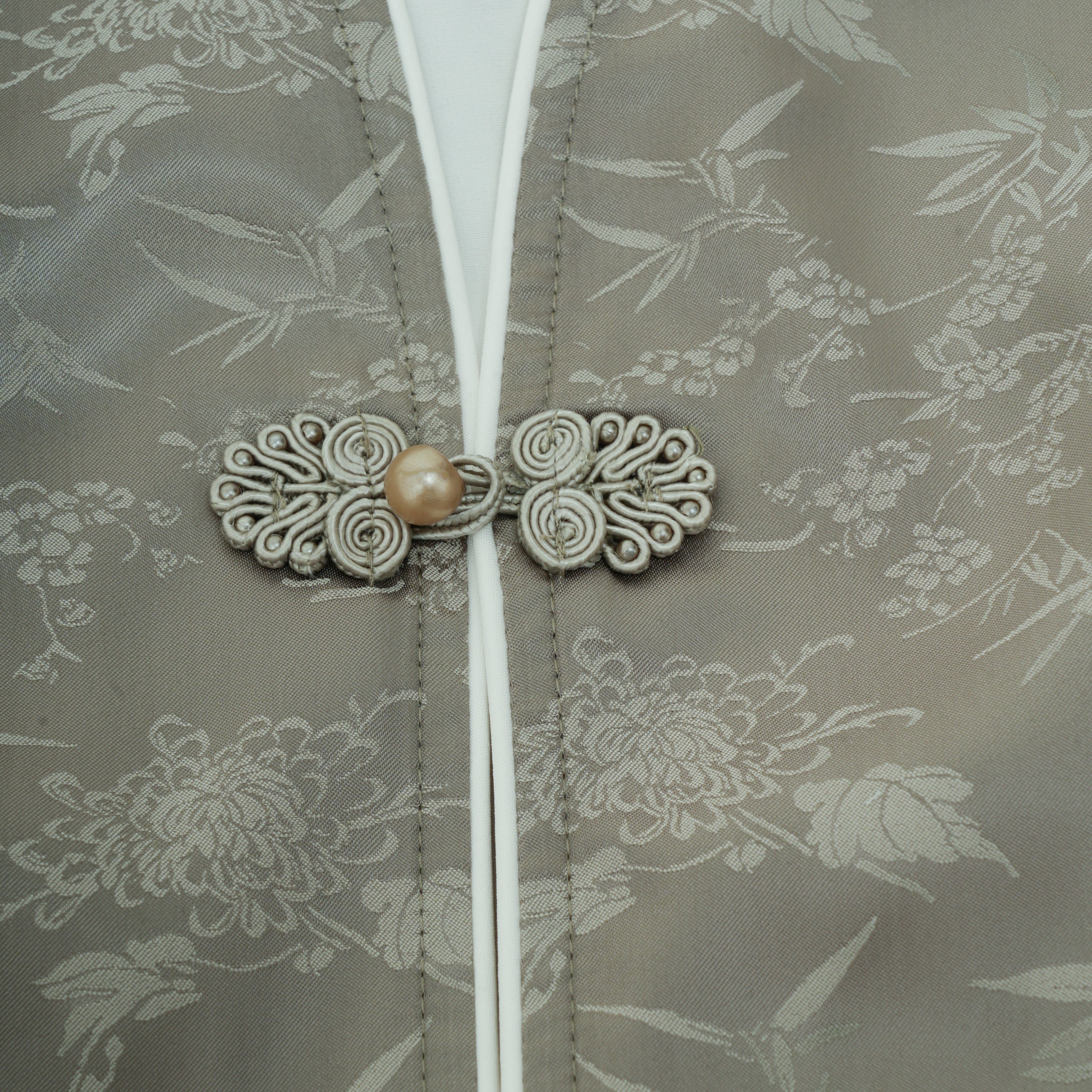 Close-up of a traditional Chinese-style garment featuring a decorative frog closure with a central pearl, set against a taupe brocade fabric with a floral pattern.